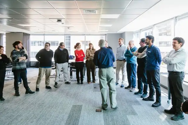 Un grupo de personas conversando de pie, en círculo, en una sala con ventanales